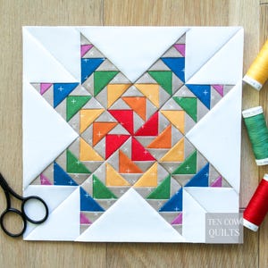 May include: A vibrant quilt block with a starburst pattern. The centre has a pinwheel design in red, orange, yellow, green, and blue. White triangles frame the design. The text "TEN COW QUILTS" is visible in the lower right corner.