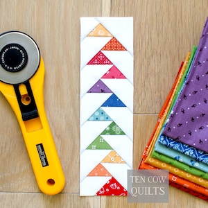 Puede incluir: Un bloque de patchwork hecho a mano con patrones triangulares coloridos en rojo, naranja, rosa, morado, azul, verde y blanco. Un cortador rotatorio amarillo y una pila de tela est&aacute;n al lado del bloque. El texto "TEN COW QUILTS" est&aacute; en la esquina inferior derecha.