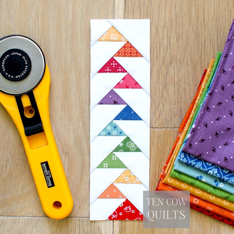 Puede incluir: Un bloque de patchwork hecho a mano con patrones triangulares coloridos en rojo, naranja, rosa, morado, azul, verde y blanco. Un cortador rotatorio amarillo y una pila de tela est&aacute;n al lado del bloque. El texto "TEN COW QUILTS" est&aacute; en la esquina inferior derecha.