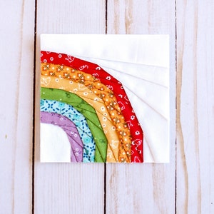 May include: A rainbow-colored fabric quilt block with a white background. The rainbow is made up of red, orange, yellow, green, blue, and purple fabric strips.
