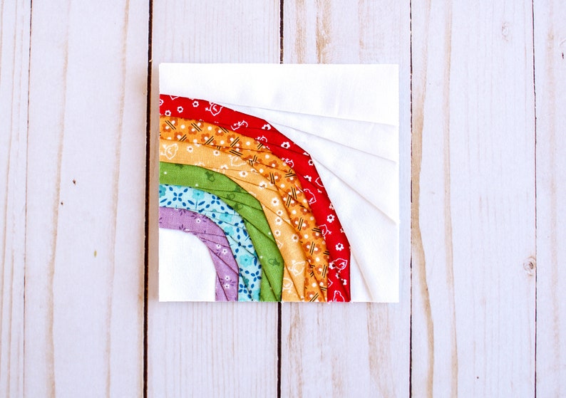 May include: A rainbow-colored fabric quilt block with a white background. The rainbow is made up of red, orange, yellow, green, blue, and purple fabric strips.