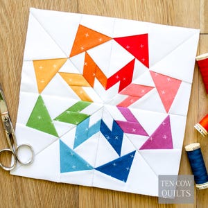 May include: A colorful quilt block with a starburst design. The block features triangles in shades of red, orange, yellow, green, blue, and purple, set against a white background. The quilt block is made of fabric and has a stitched pattern.