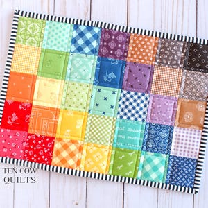 May include: A rainbow patchwork quilt with a black and white striped border. The quilt is made of various colourful fabrics with different patterns. The quilt is labelled "TEN COW QUILTS".