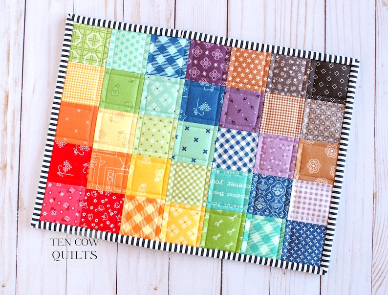 May include: A rainbow patchwork quilt with a black and white striped border. The quilt is made of various colourful fabrics with different patterns. The quilt is labelled "TEN COW QUILTS".