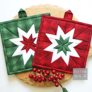 Puede incluir: Dos agarraderas cuadradas acolchadas con un patrón de estrella de ocho puntas en el centro. Una es de cuadros verdes con una estrella roja y blanca, y la otra es de cuadros rojos con una estrella verde y blanca. Ambas tienen una trabilla.