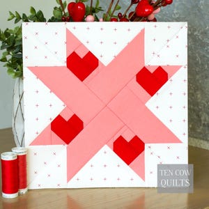 Könnte beinhalten: Ein quadratischer Quiltblock mit einem rosa Sterndesign und vier roten Herzakzenten. Der Hintergrund ist weiß mit kleinen roten Kreuzen. Der Text "TEN COW QUILTS" ist sichtbar. Zwei rote Garnrollen befinden sich im Vordergrund. Valentinstag Dekoration.