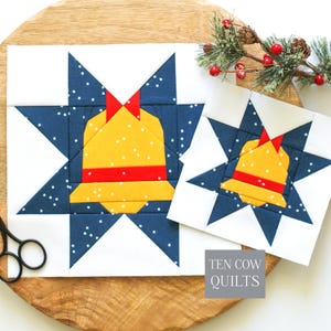 May include: Two square quilt blocks featuring a central yellow bell with a red band and bow, set against a dark blue star background with white speckles. The blocks are white-bordered and include the text TEN COW QUILTS in a gray box.