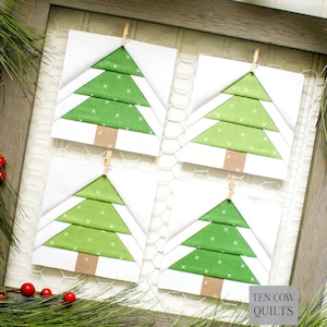 Pode incluir: Quatro blocos de colcha de árvore de Natal, cada um com um desenho de árvore verde sobre um fundo branco. As árvores têm troncos castanhos e são presas a uma tela de arame com pequenos prendedores de roupa de madeira. O texto "TEN COW QUILTS" é visível.