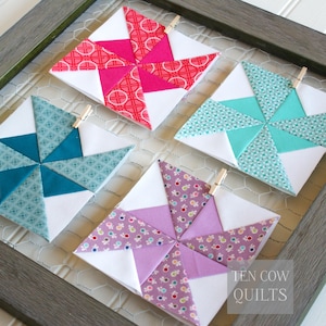 Pode incluir: Quatro blocos de colcha coloridos e feitos &agrave; m&atilde;o em uma moldura cinza. Cada bloco apresenta um design de cata-vento com tecidos brancos, turquesa, rosa e roxo. Os blocos s&atilde;o presos com pequenos prendedores de roupa de madeira. O texto "TEN COW QUILTS" &eacute; vis&iacute;vel.