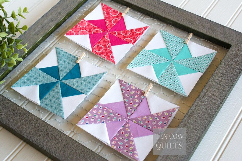Pode incluir: Quatro blocos de colcha coloridos e feitos &agrave; m&atilde;o em uma moldura cinza. Cada bloco apresenta um design de cata-vento com tecidos brancos, turquesa, rosa e roxo. Os blocos s&atilde;o presos com pequenos prendedores de roupa de madeira. O texto "TEN COW QUILTS" &eacute; vis&iacute;vel.