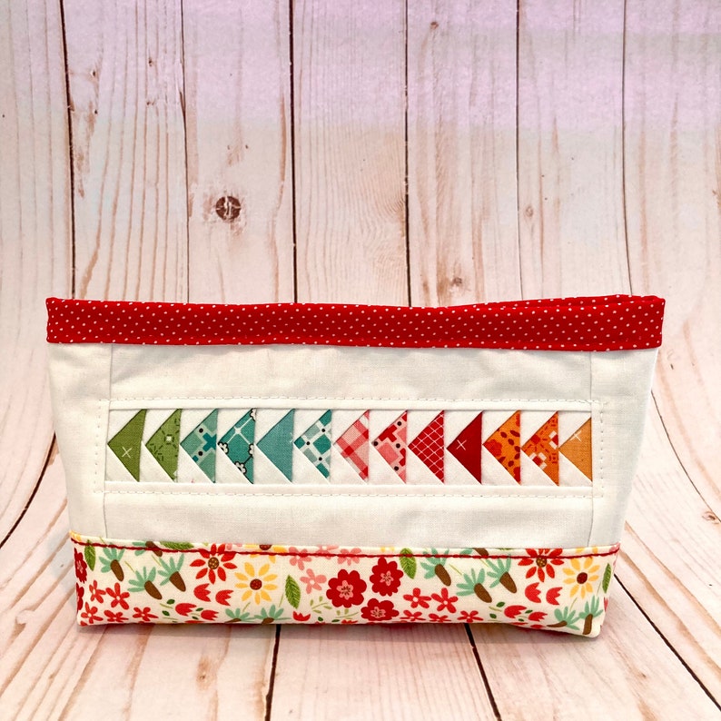 Puede incluir: Una cesta de almacenamiento de tela blanca con un borde rojo de lunares y un estampado floral en la parte inferior. La cesta tiene una fila de tri&aacute;ngulos de patchwork coloridos en la parte delantera.