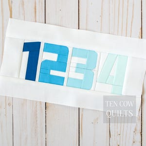 May include: A rectangular fabric quilt block featuring the numbers 1, 2, 3, and 4 pieced together in a gradient of blue shades, ranging from dark navy to light mint, set against a solid white background with the text TEN COW QUILTS.