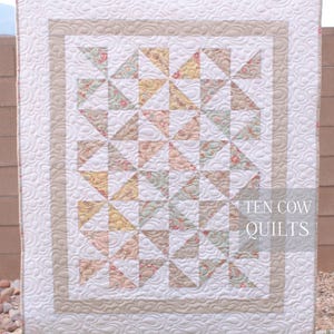 May include: A white quilt with a beige border and a pattern of pink, yellow, and green floral fabric triangles. The quilt is quilted with a wavy pattern. The text "TEN COW QUILTS" is in the bottom right corner.