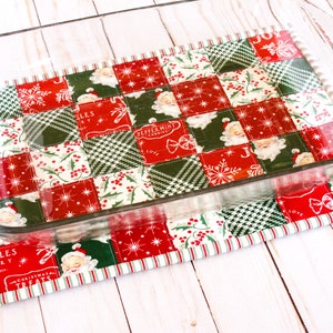 May include: A red and green Christmas-themed quilted hot pad with a glass baking dish on top. The hot pad is made of patchwork fabric with various holiday designs, including snowflakes, holly, and Santa Claus.