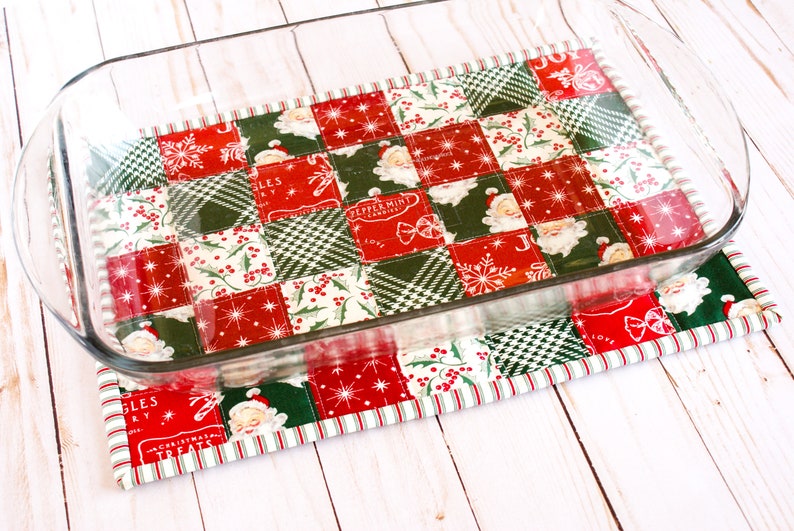May include: A red and green Christmas-themed quilted hot pad with a glass baking dish on top. The hot pad is made of patchwork fabric with various holiday designs, including snowflakes, holly, and Santa Claus.