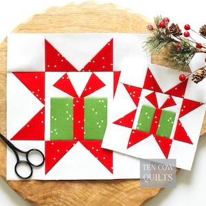 May include: Two square quilt blocks featuring a central green gift box with a red ribbon, surrounded by a red and white star pattern. The fabric has a subtle white speckled texture. Black scissors are positioned on the left side of the blocks.