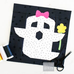 Op de afbeelding: Een handgemaakt quiltblok met een spookontwerp. De geest is wit met zwarte stippen, een roze strik en een gele bloem. De achtergrond is zwart met witte sterren. Het blok is vierkant en heeft de tekst "TEN COW QUILTS".