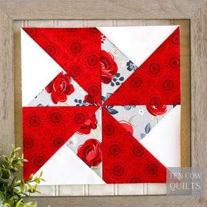 Pode incluir: Bloco de colcha emoldurado com um design de cata-vento. O bloco de colcha &eacute; composto por tecido floral vermelho, tecido branco e tecido floral azul claro. A moldura &eacute; de cor cinza claro. O texto "TEN COW QUILTS" &eacute; vis&iacute;vel no canto inferior direito.