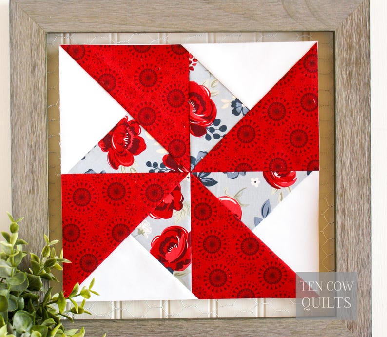 Pode incluir: Bloco de colcha emoldurado com um design de cata-vento. O bloco de colcha &eacute; composto por tecido floral vermelho, tecido branco e tecido floral azul claro. A moldura &eacute; de cor cinza claro. O texto "TEN COW QUILTS" &eacute; vis&iacute;vel no canto inferior direito.
