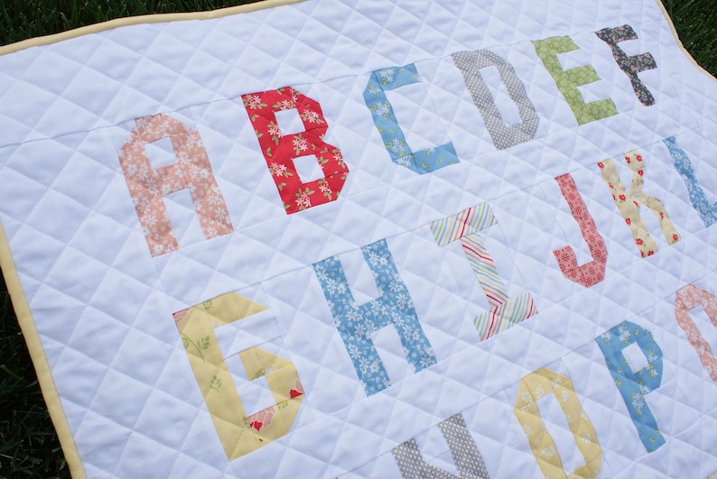 May include: A white quilt with the alphabet in colorful patterned fabric letters. The letters are sewn onto the quilt in a staggered pattern.