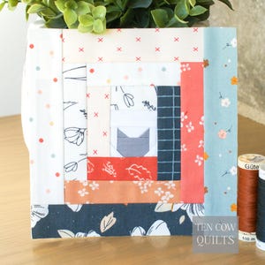 May include: A square fabric block quilt with a cat design in the centre. The quilt features various patterned fabrics in white, red, blue, and orange. The text "TEN COW QUILTS" is visible on the bottom right.