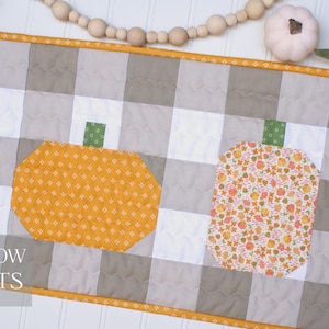 May include: A quilted table runner with a gray and white checkered pattern. The runner features appliqued pumpkins in orange and floral patterns with green stems. The text "TEN COW QUILTS" is visible.