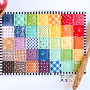 May include: A colourful patchwork quilt hot pad with a rainbow pattern. The hot pad is made of various fabrics with different patterns, including gingham, polka dots, and floral prints. The hot pad is edged with a black and white striped border. The text "TEN COW QUILTS" is printed on the bottom of the hot pad.