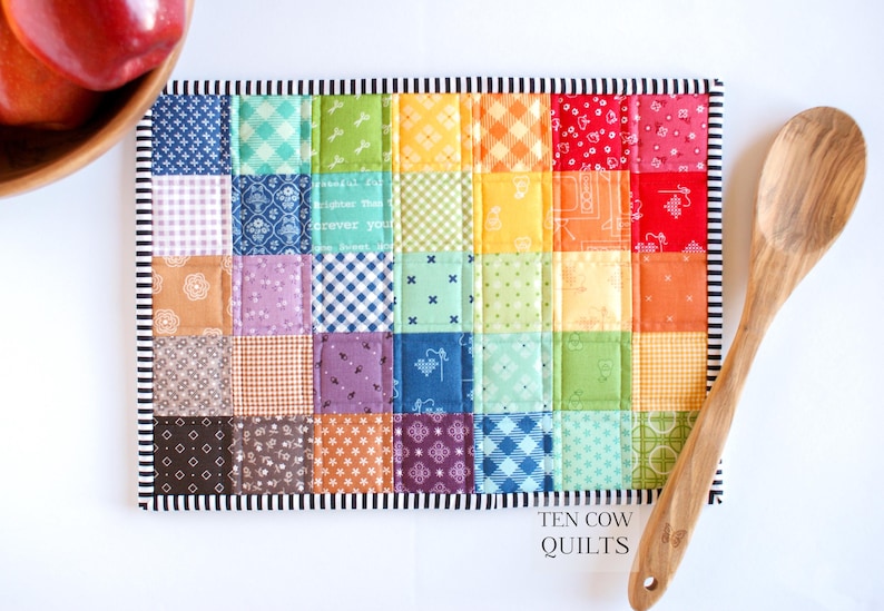 May include: A colourful patchwork quilt hot pad with a rainbow pattern. The hot pad is made of various fabrics with different patterns, including gingham, polka dots, and floral prints. The hot pad is edged with a black and white striped border. The text "TEN COW QUILTS" is printed on the bottom of the hot pad.