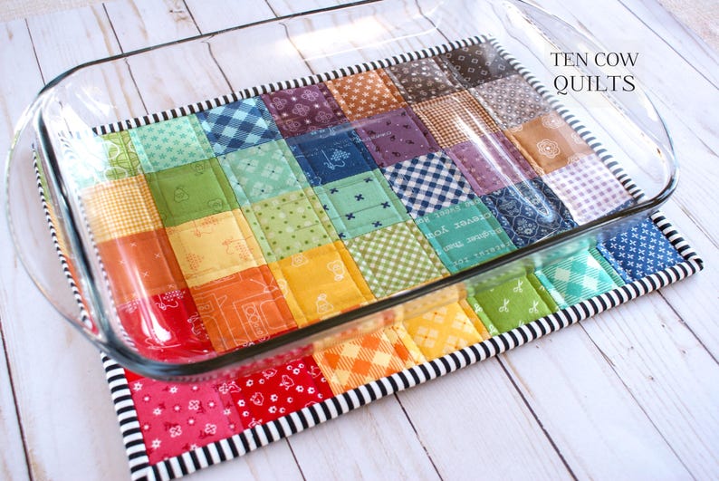 May include: A colourful patchwork quilt hot pad with a black and white trim, designed to protect surfaces from hot dishes. The hot pad is placed on top of a clear glass baking dish. The text "TEN COW QUILTS" is visible in the top right corner of the image.
