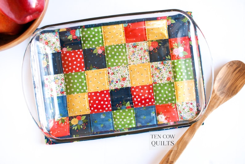 May include: A glass baking dish with a colourful patchwork fabric hot pad underneath. The fabric is made of squares in shades of red, yellow, green, blue, and white. The text "TEN COW QUILTS" is visible in the bottom right corner of the image.