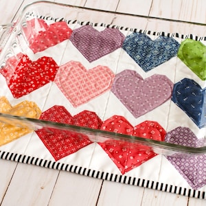 May include: A glass baking dish sits on a quilted hot pad with a chevron pattern of colorful hearts. The hot pad has a black and white striped border.