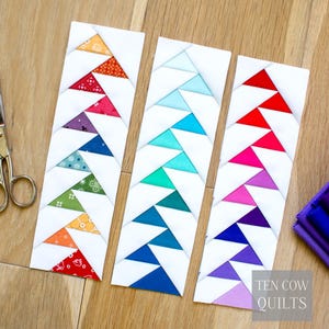 Puede incluir: Tres marcap&aacute;ginas de tela blanca con patrones triangulares coloridos. Cada marcap&aacute;ginas presenta una combinaci&oacute;n de colores diferente: rojo, naranja, amarillo, verde, azul y morado. El texto "TEN COW QUILTS" es visible en la esquina inferior derecha.