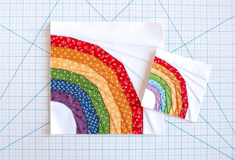 May include: Two fabric squares with a rainbow design. The larger square is white with a rainbow of fabric strips sewn in a curved shape. The smaller square is white with a smaller rainbow design.