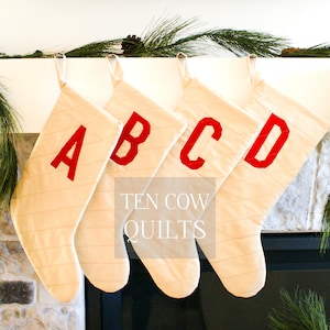 May include: Four white Christmas stockings with red felt letters spelling out "ABCD" hanging on a white mantel. The stockings are lined up in a row and have a subtle striped pattern. The text "TEN COW QUILTS" is printed below the stockings.