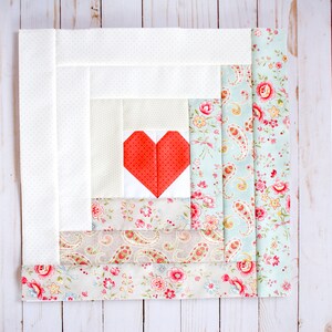 Puede incluir: Un bloque de colcha blanco y floral con un parche en forma de coraz&oacute;n rojo cosido en el centro. El bloque est&aacute; formado por varias capas de tela, cada una con un patr&oacute;n diferente.