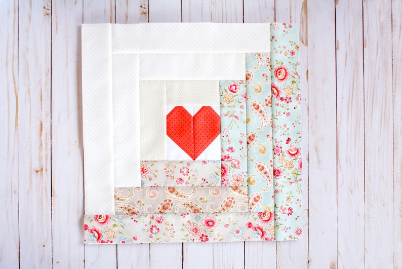 Puede incluir: Un bloque de colcha blanco y floral con un parche en forma de coraz&oacute;n rojo cosido en el centro. El bloque est&aacute; formado por varias capas de tela, cada una con un patr&oacute;n diferente.