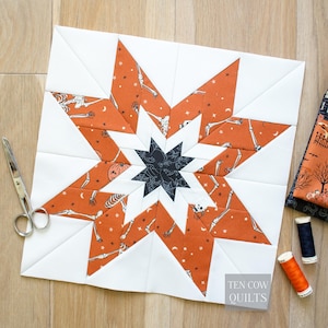 Puede incluir: Un bloque de edredón con temática de Halloween con un diseño de estrella. Las puntas de la estrella son naranjas con un estampado de esqueleto, sobre un fondo blanco. Una estrella azul oscuro está en el centro. El texto "TEN COW QUILTS" está en la esquina inferior derecha.