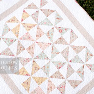 May include: A white quilt with a beige border and a pattern of floral print triangles. The quilt is quilted with a swirl pattern. The text "TEN COW QUILTS" is visible in the lower left corner.
