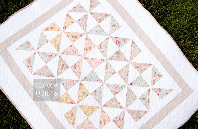 May include: A white quilt with a beige border and a pattern of floral print triangles. The quilt is quilted with a swirl pattern. The text "TEN COW QUILTS" is visible in the lower left corner.