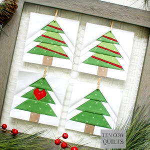 Pode incluir: Quatro blocos quadrados de colcha de árvore de Natal, cada um com um design de árvore verde, detalhes vermelhos e um tronco marrom. Um bloco apresenta um coração vermelho. Os blocos são presos a um fundo de malha de arame dentro de uma moldura de madeira. O texto "TEN COW QUILTS" é visível.