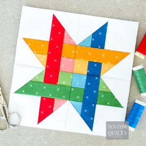 Puede incluir: Un bloque de patchwork colorido con un diseño de estrella. La estrella está compuesta por piezas de tela rojas, naranjas, rosas, verdes y azules, cada una con detalles de puntadas de cruz blancas, sobre un fondo blanco. La imagen incluye carretes de hilo y tijeras.