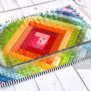 May include: A colorful patchwork quilt hot pad with a black and white trim, designed to protect surfaces from hot dishes. The hot pad is made with a variety of fabrics in shades of red, orange, yellow, green, blue, and purple. The hot pad is placed under a clear glass baking dish.