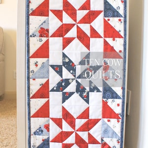 以下が含まれることがあります： 赤、白、青の星柄のキルトテーブルランナー。ランナーは白いキャビネットに掛けられており、中央には「TEN COW QUILTS」の文字が書かれています。