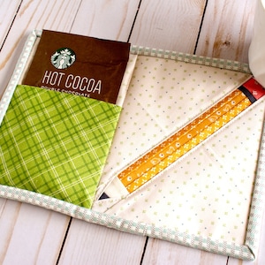 May include: A green and white checkered fabric coaster with a brown packet of hot cocoa that says "Hot Cocoa Double Chocolate" and a yellow pencil design.