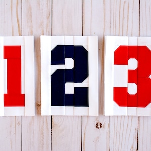 Puede incluir: Tres números, 1, 2 y 3, se muestran en un fondo de madera blanco. Los números están hechos de tela y son rojo, azul y rojo, respectivamente.