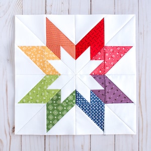May include: A colourful patchwork quilt block featuring a star design with eight points. The star is made up of eight triangles in different colours, including red, orange, yellow, green, blue, purple, and pink. The background of the block is white.