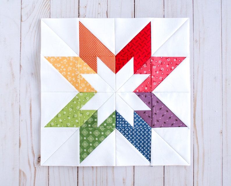 May include: A colourful patchwork quilt block featuring a star design with eight points. The star is made up of eight triangles in different colours, including red, orange, yellow, green, blue, purple, and pink. The background of the block is white.