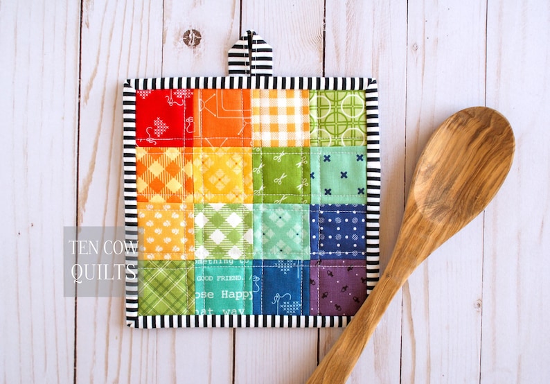 May include: A square quilted potholder featuring a patchwork grid of multicolored fabric squares in red, orange, yellow, green, blue, and purple with various patterns. It has a black and white striped border and hanging loop. A wooden spoon is placed beside it.