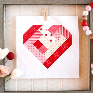 May include: A white fabric square with a red, pink, and white patchwork heart design. The heart is made of various patterned fabrics, including stripes, polka dots, and hearts.