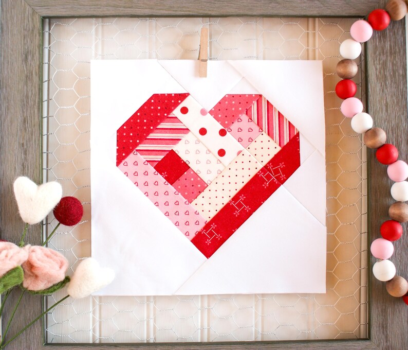 May include: A white fabric square with a red, pink, and white patchwork heart design. The heart is made of various patterned fabrics, including stripes, polka dots, and hearts.